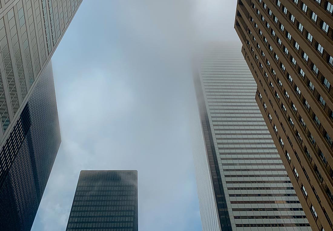 Dramatic, low-angle view of multiple modern skyscrapers partially shrouded in fog, emphasizing the massive scale and imposing presence of core urban real estate.