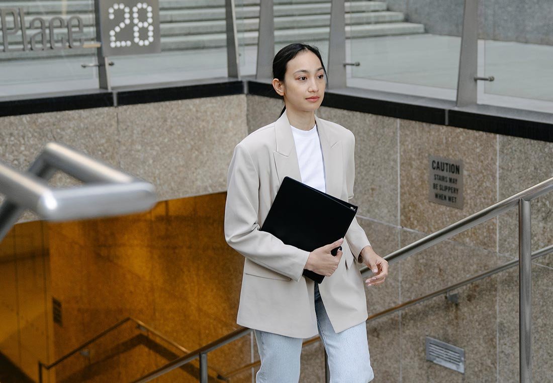 A focused professional woman in a blazer holds a black folder while descending a modern staircase, symbolizing action and corporate readiness in an urban setting.